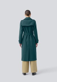 Long teal wool coat with a belted waist, notched lapels, and buttoned cuffs. Back vent for movement, worn over beige wide-leg trousers.