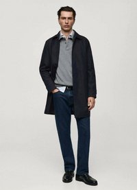 Herroutfit som har en lång svart kappa, en grå polo, en vit skjorta under, mörkblå jeans och svarta skor. Alla plagg är i grundläggande, enfärgade nyanser.