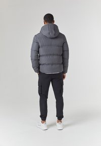 Grå puffjakke med hette, med horisontale sømmer, kombinert med svarte cargo-bukser og hvite sneakers, sett fra baksiden.