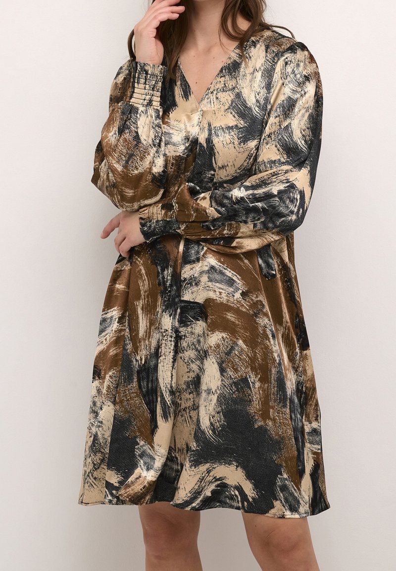 Femme portant une robe mi-longue à manches longues avec un motif abstrait composé de coups de pinceau marron, beige et noir, debout devant un fond uni.