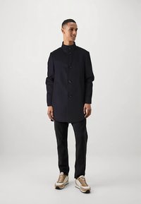 Cappotto blu navy con colletto alto, chiusura con bottoni e tasche laterali, abbinato a pantaloni neri e sneakers beige. Tessuto dalla texture liscia.