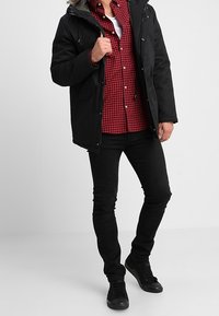 Czarna kurtka puchowa z kapturem wykończonym futrem, noszona na czerwonym koszulowym T-shircie w kratę, zestawiona z czarnymi dżinsami i czarnymi sneakersami.
