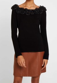 Svart långärmad topp med volangig genomskinlig halsringning, i kombination med en brun läderskjol. Slät yta, åtsittande design och minimal detaljering.