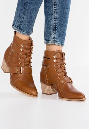 Botas de tobillo de imitación de cuero marrón con punta afilada, cordones en la parte delantera y una correa con hebilla en el lateral. Presenta un tacón apilado y un diseño con cortes.