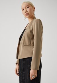 Beige cropped blazer with a structured design, featuring a single front closure, long sleeves, and a smooth, matte texture.