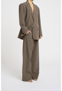 Blazer marron sur mesure avec un col en V profond et des clous décoratifs sur les épaules, associé à un pantalon à jambes larges avec des détails de clous sur les côtés.