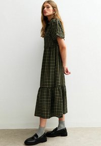 Robe maxi à carreaux verts avec de courtes manches bouffantes, une taille cintrée et une jupe à volants. Associée à des chaussures noires et des chaussettes grises.