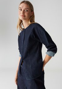 Dunkelblaues Jeanshemd mit rundem Ausschnitt und Knopfleiste. Mit hochgekrempelten Ärmeln aus hellerer Jeans. Figurbetont und leicht leger geschnitten.