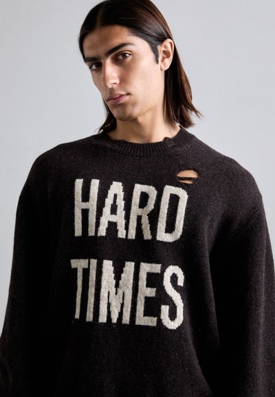 Dunkelbrauner gestrickter Pullover mit dem weißen Schriftzug "HARD TIMES" und einem kleinen Loch im Stoff an der linken Schulter.