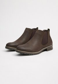Un par de botas Chelsea de cuero marrón con paneles laterales elásticos y tacones bajos apilados sobre un fondo blanco liso.