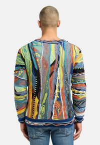 Multifarvet strikket sweater med abstrakte mønstre og strukturerede detaljer; har lange ærmer og en rund hals, parret med blå jeans.