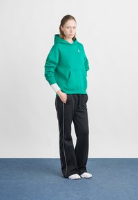 Πράσινο φούτερ με κουκούλα και τσέπη καγκουρό; μαύρο παντελόνι με λευκές λωρίδες στο πλάι; το μοντέλο στέκεται σε ένα μπλε χαλί μπροστά από ένα λευκό φόντο.