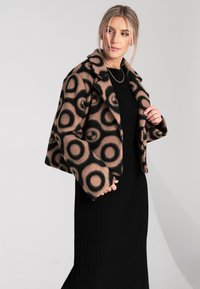 Brązowa i czarna kurtka w wzory z wyciętym kołnierzem, z okrągłymi wzorami, noszona na czarnej, wżeranej sukience. Miękka faktura, luźny krój.
