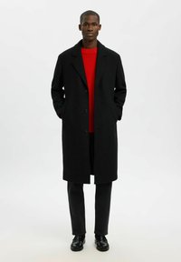 Mann in einem langen schwarzen Mantel über einem roten Pullover, schwarzen Hosen und schwarzen Slippern, der mit Händen in den Manteltaschen vor einem einfachen Hintergrund steht.