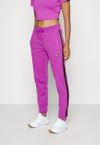 Fuchsia joggers with drawstring waist, side pockets, and black side stripes. Paired with white sneakers, featuring subtle logo accents.