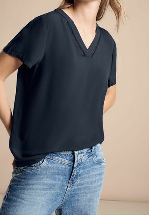 Femme portant un chemisier bleu marine foncé à col en V et manches courtes, et un jean en denim bleu clair, debout avec les mains dans le dos sur un fond beige.