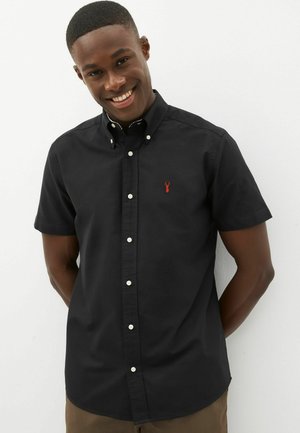 Camisa negra de manga corta con cuello abotonado, que presenta un logo de langosta roja en el pecho y botones blancos. Textura de tela suave.
