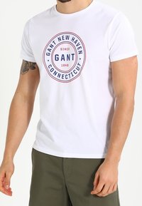 Camiseta de algodón blanca con un estampado gráfico redondo que dice "GANT NEW HAVEN CONNECTICUT" en azul marino y rojo. Mangas cortas y cuello redondo.