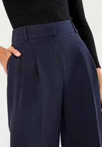 Navy pleated trousers made from textured fabric, featuring a high waistband and side pockets, paired with a fitted black ribbed top.