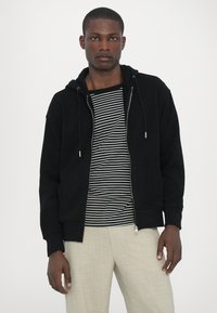 Felpa nera con zip e coulisse, tasche, indossata sopra una maglietta a righe bianche e nere. Il modello indossa pantaloni chiari.