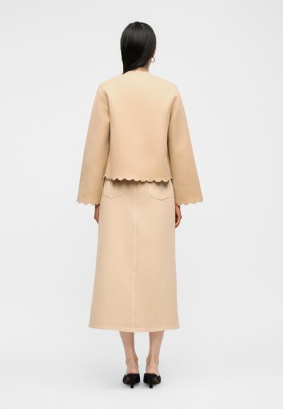 Vrouw met lang zwart haar, gekleed in een beige jas met gegolfde rand en een bijpassende lange rok, staand op blote voeten tegen een witte achtergrond.