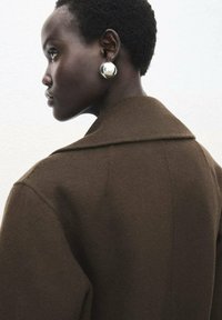 Abrigo de lana marrón con un amplio cuello, con una textura suave. Un gran pendientes de plata brillante complementa el look.