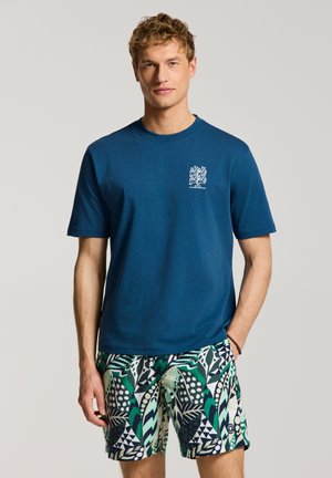 Mand iført en blå t-shirt med lille hvid grafik og grønne, sorte og hvide mønstrede shorts, stående mod en ensfarvet grå baggrund.