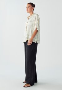 Blusa ligera beige con líneas geométricas negras, diseño de botones y mangas cortas, combinada con pantalones amplios de textura negra.