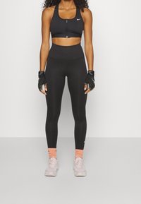 Czarny biustonosz sportowy na zamek z przodu, czarne legginsy z wysokim stanem, czarne rękawiczki bezpalcowe; białe logo Nike; jasnoróżowe sneakersy; pomarańczowe skarpetki.