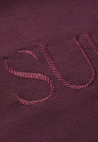La tela burdeos presenta un bordado en relieve de las letras "SU". La textura es suave con un ligero brillo. El patrón es sencillo y limpio.
