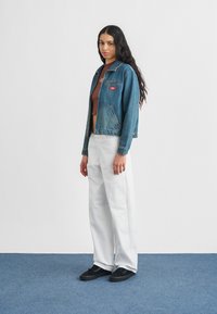 Γυναίκα με μακριά σκουρόχρωμα μαλλιά που φοράει κολλητή καφέ μπλούζα, μπλε τζιν μπουφάν, λευκό παντελόνι και μαύρα αθλητικά παπούτσια, στέκεται σε μπλε χαλί.