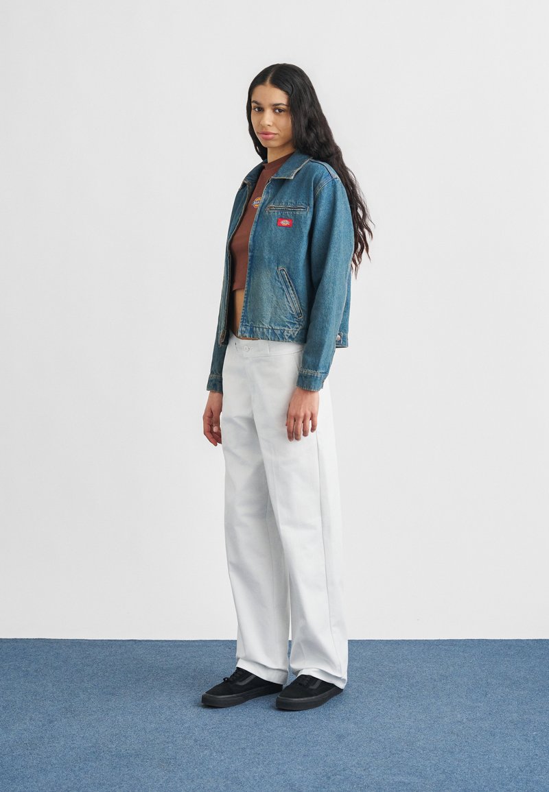 Vrouw met lang donker haar, gekleed in een cropped bruin topje, een blauwe spijkerjas, witte broek en zwarte sneakers, staand op een blauw tapijt.