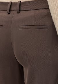 Gros plan sur un pantalon marron tailleur avec poches passepoilées à l'arrière et passants de ceinture, porté avec un pull côtelé beige.