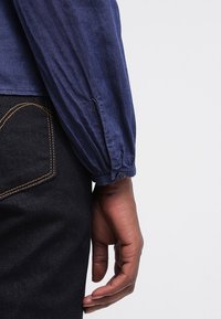 Mörkhyad hand avslappnad vid sidan, iklädd en blå långärmad skjorta med knäppta manschetter och svarta byxor med synlig bakfickssöm.