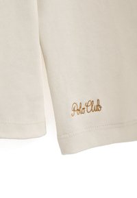 Cremefarbenes Polo-Shirt mit geripptem Muster, ausgestattet mit dem goldgestickten "Polo Club"-Logo am Saum. Kurze Ärmel.