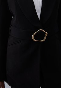 Zwarte blazer met een gladde textuur, voorzien van een brede riem en een gouden ovale gesp. Heeft zakdetails en een getailleerde pasvorm.