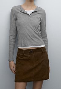 Maglietta grigia a maniche lunghe con scollatura henley e tre bottoni; abbinata a una mini gonna in suede marrone con chiusura frontale a bottoni.