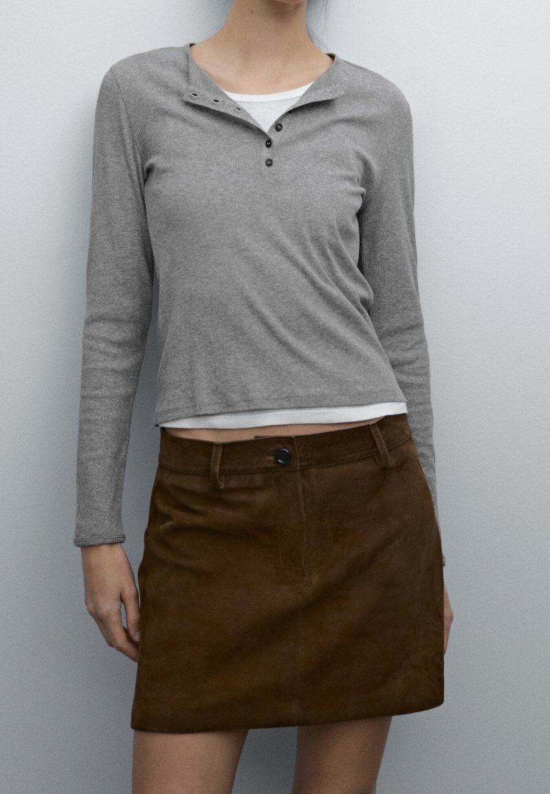 Maglietta grigia a maniche lunghe con scollatura henley e tre bottoni; abbinata a una mini gonna in suede marrone con chiusura frontale a bottoni.