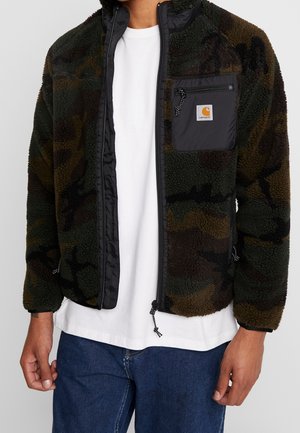 Personne portant une veste polaire camouflage verte et marron avec une poche zippée noire, sur un t-shirt blanc et un jean bleu, debout les mains détendues.