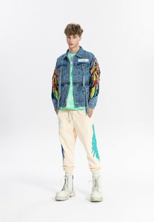 Joven con chaqueta vaquera con alas de lentejuelas de colores en las mangas, camiseta verde tie-dye, pantalones deportivos crema con estampados de alas azules y botas blancas.