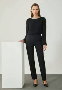 Blusa negra con mangas transparentes y cuello adornado, combinada con pantalones negros ajustados y tacones negros de punta, posando contra un pedestal blanco.