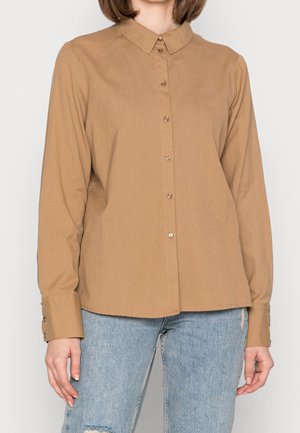 Femme portant une chemise marron camel à manches longues, boutonnée avec un col, et un jean bleu clair déchiré.
