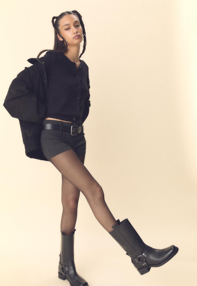 Črn cropped top s gumbi, črne prugaste kratke hlače, prosojne črne nogavice in črne visoke čevlje z zaponkami ter teksturiranimi detajli.