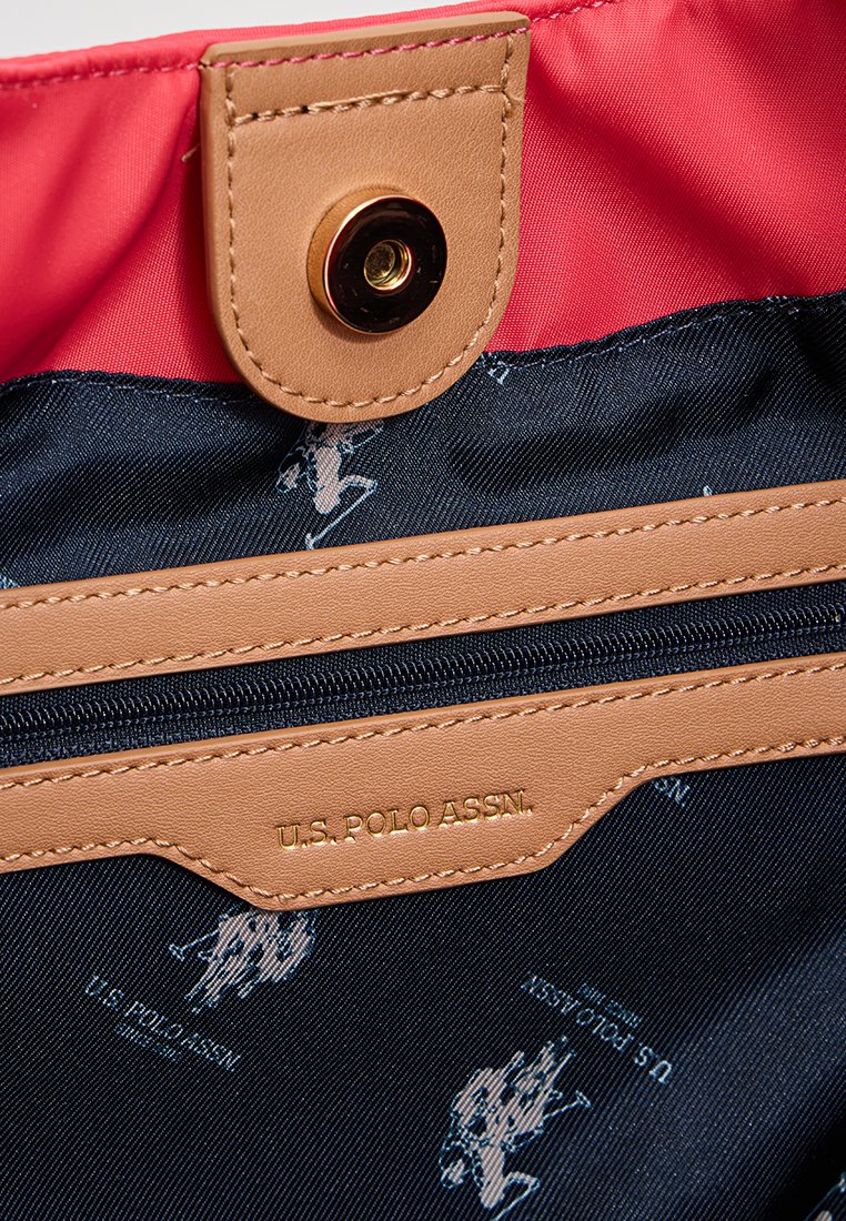 Brązowe skórzane akcenty z zapięciem na zatrzask, czerwone wykończenie oraz granatowe wnętrze zdobione powtarzającym się wzorem logo U.S. Polo Assn. W komplecie kieszeń na zamek.