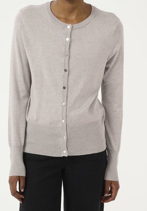 Hellgrauer, geknöpfter Cardigan, getragen von einer Person mit dunkler Hose, zeigt Oberkörper und Arme vor einfarbigem Hintergrund.