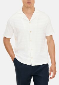 Camicia bianca a maniche corte con un design testurizzato, dotata di un colletto alla francese e bottoni marroni chiari, abbinata a pantaloni blu scuro.