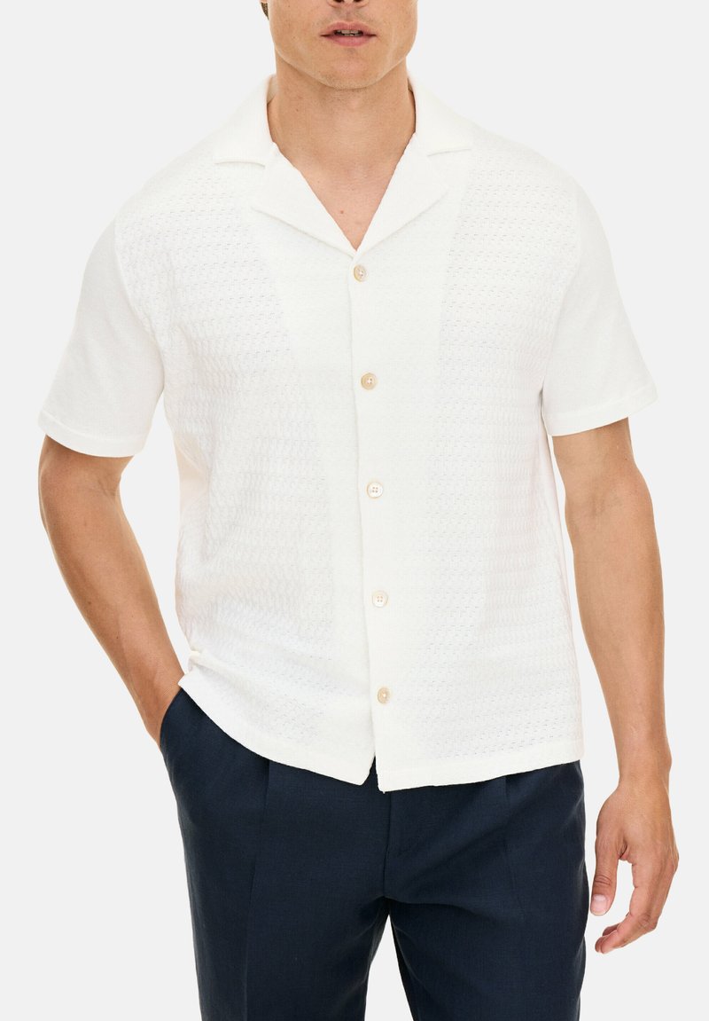 Camicia bianca a maniche corte con un design testurizzato, dotata di un colletto alla francese e bottoni marroni chiari, abbinata a pantaloni blu scuro.