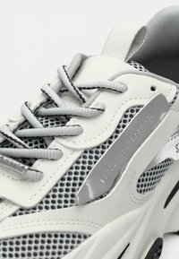 Baskets blanches avec des accents gris, dotées de panneaux en mesh, de lacets ronds et d'un détail latéral réfléchissant. Semelle structurée et col rembourré.