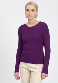 Lila gerippter Strickpullover mit langen Ärmeln und rundem Halsausschnitt, kombiniert mit cremefarbenen Hosen. Glatte Textur und taillierter Schnitt.