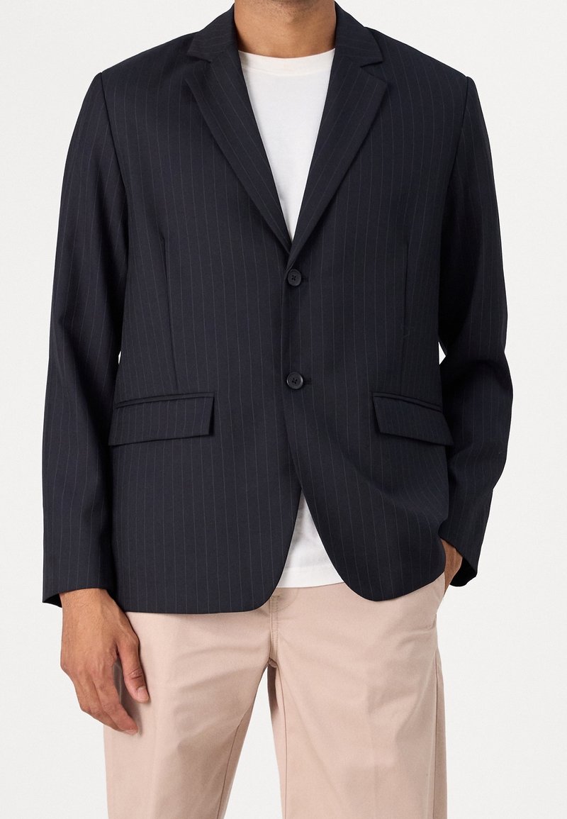 Homme portant un blazer foncé à fines rayures sur un t-shirt blanc et un pantalon beige, debout avec une main dans la poche devant un fond uni.